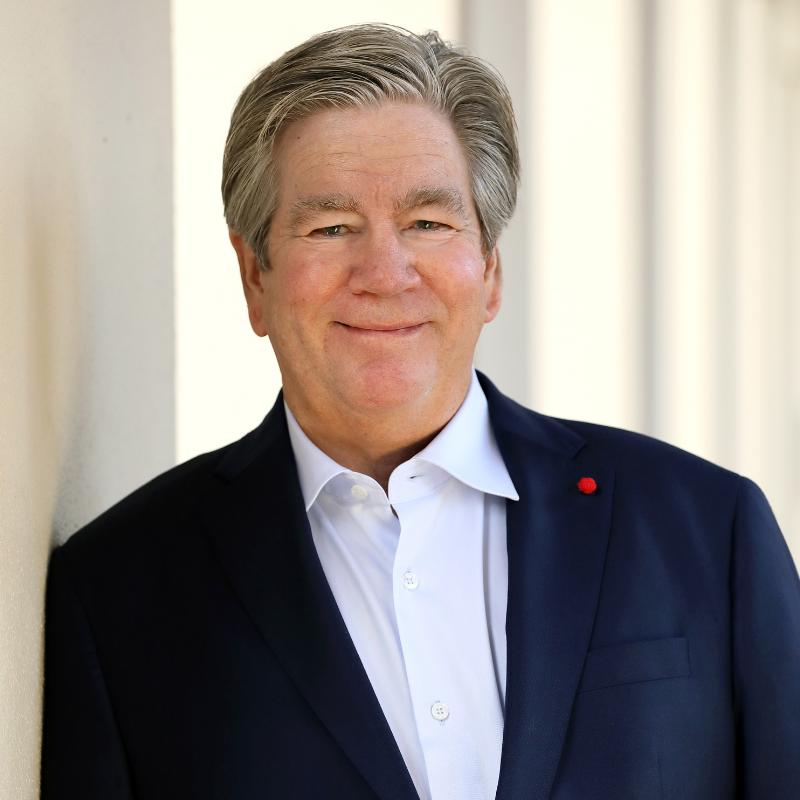 A smiling older man with gray hair, wearing a navy blue suit jacket over a white dress shirt, stands against a light-colored wall with a blurred background.