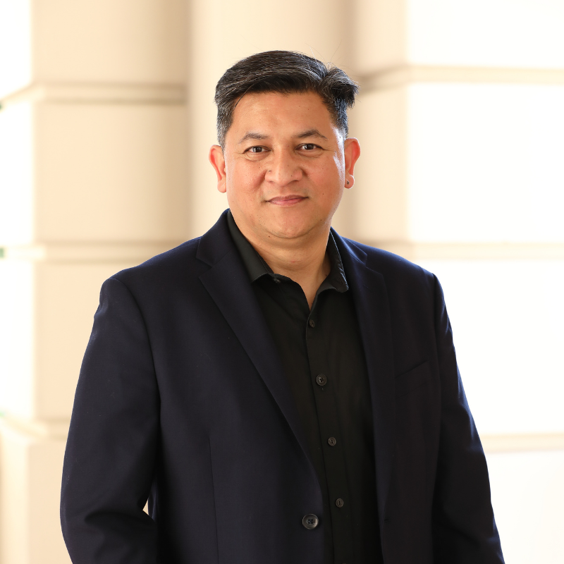 A man with short dark hair wearing a black suit jacket and black shirt stands indoors, smiling softly at the camera. The background features cream-colored walls with soft natural light.