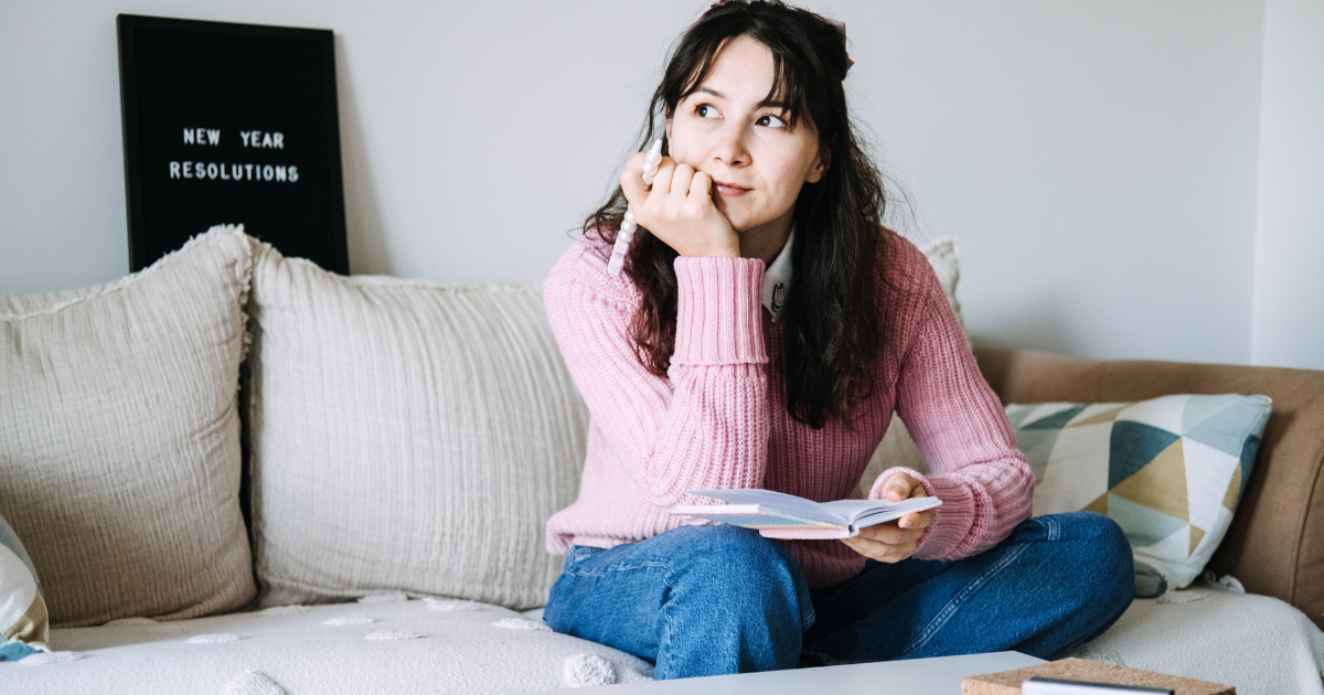 A woman sits on a couch holding a notebook and pen, looking thoughtful. Behind her, a sign says New Year Resolutions. She wears a pink sweater and blue jeans, with neutral-toned cushions in the background.