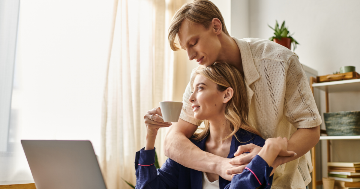 A man gently hugs a woman from behind as she sits at a desk holding a white mug and looks at her laptop, highlighting the positive impacts of relationships on mental health. Sunlight streams through the window, with houseplants in the background.