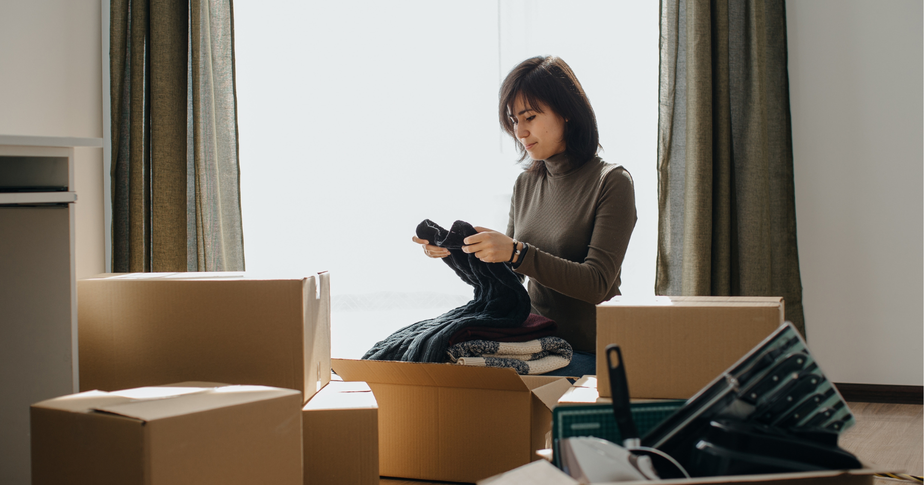 A woman sits in a bright room surrounded by cardboard boxes, packing or unpacking clothes. She holds a folded sweater and wears a turtleneck sweater, with sunlight streaming in through large windows and green curtains.