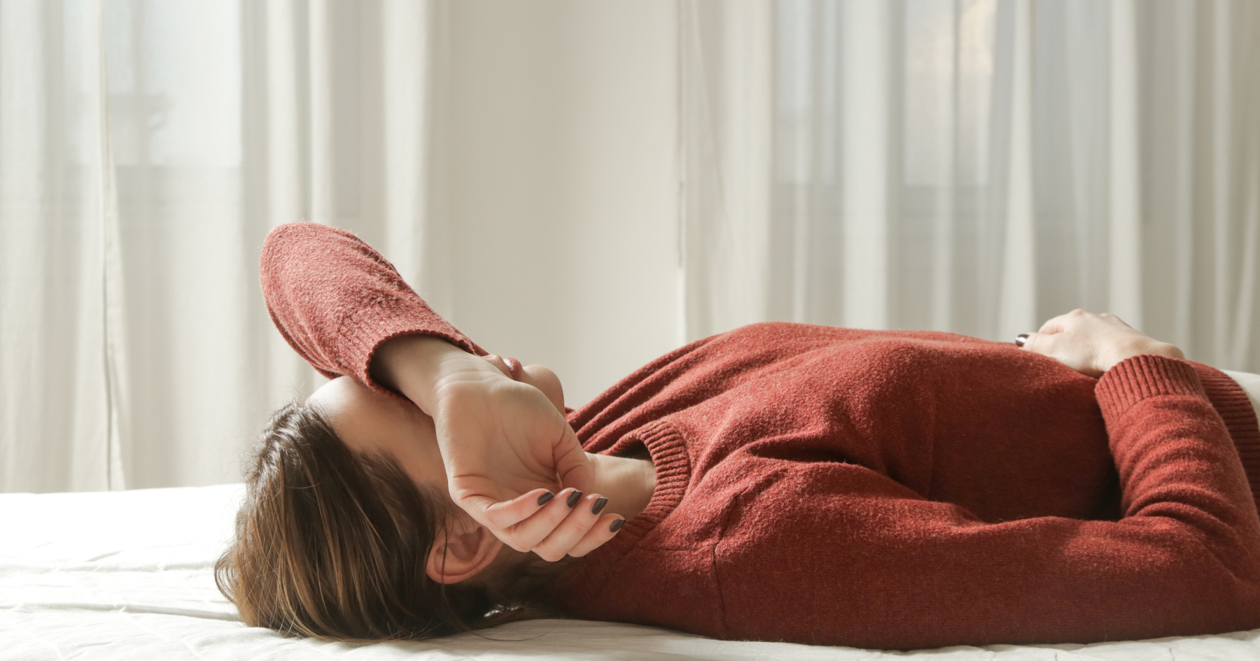 A person wearing a rust-colored sweater lies on a bed with one arm resting across their forehead, appearing tired or pensive. Soft natural light filters through sheer curtains in the background.