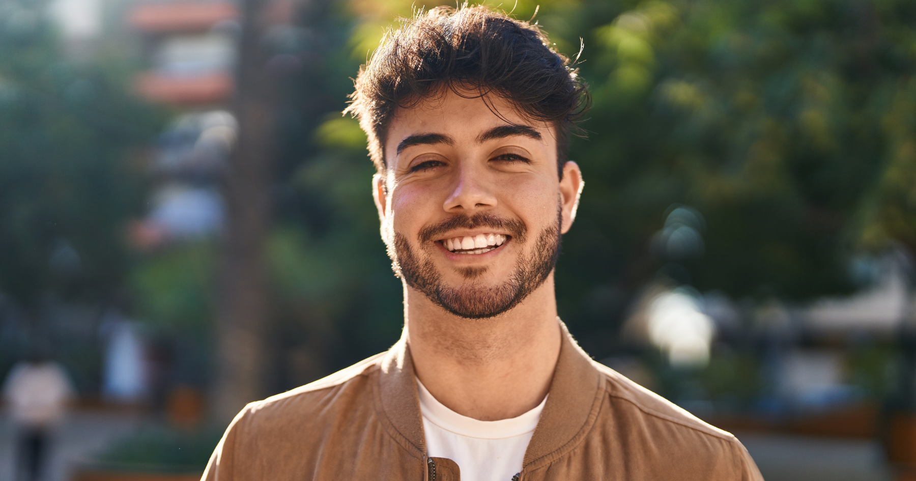 A young man with short dark hair and a beard is smiling outdoors. He is wearing a tan jacket over a white shirt, with blurred greenery and buildings in the background on a sunny day.