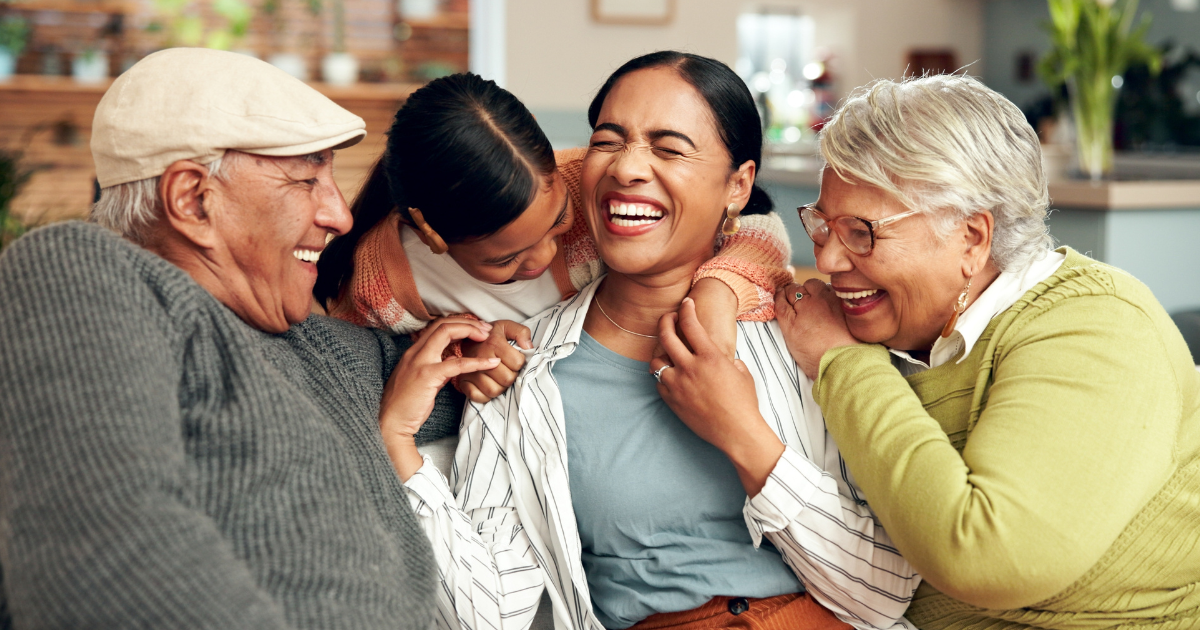 Four people, including a young girl, two older adults, and a middle-aged woman, sit closely together on a couch, laughing and smiling—a true example of building support systems for mental health in a warm, cozy home setting.