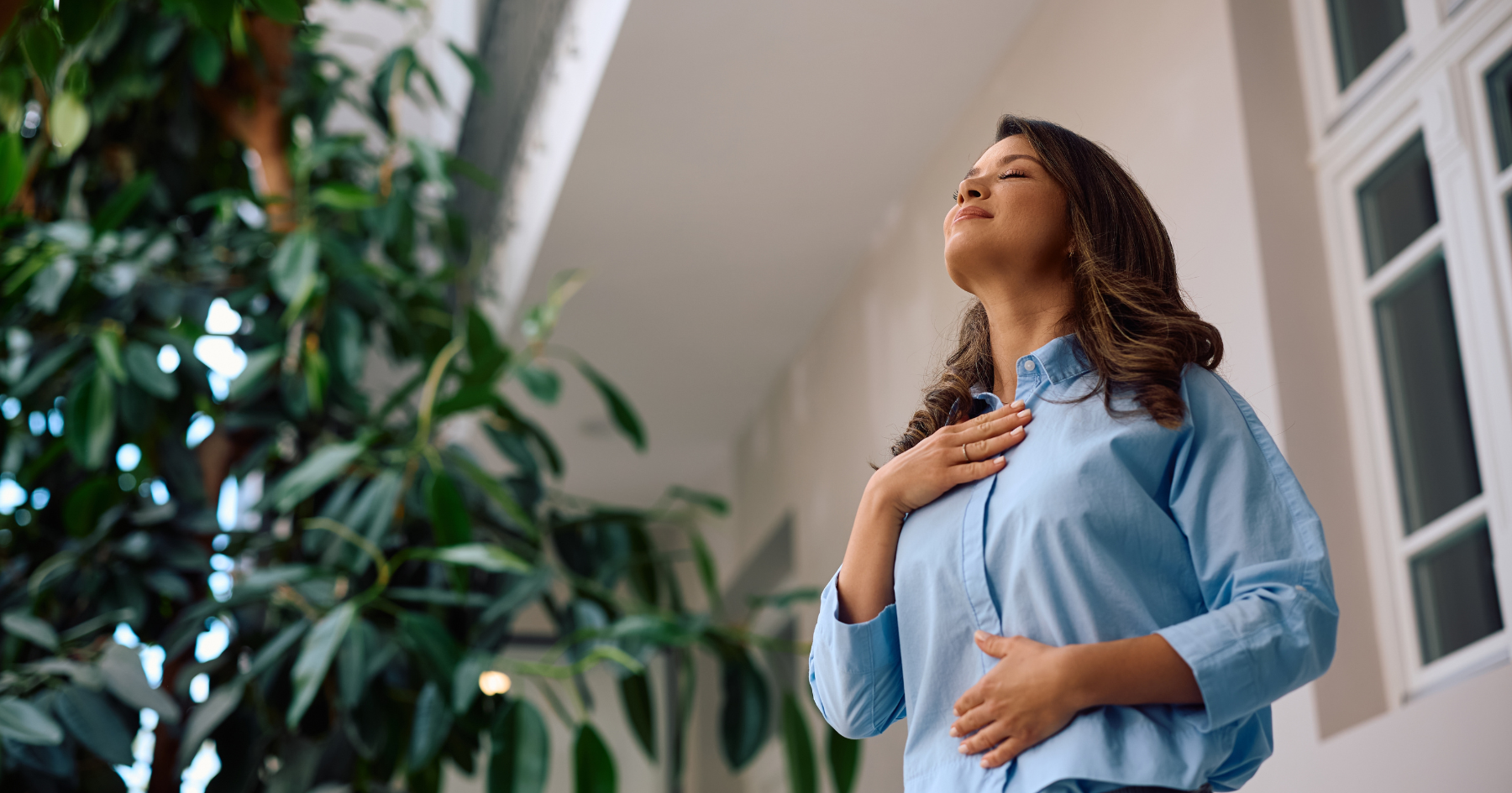 A woman stands indoors near a large plant with her eyes closed, one hand on her chest and the other on her abdomen, appearing relaxed and calm, as if practicing deep breathing or mindfulness.