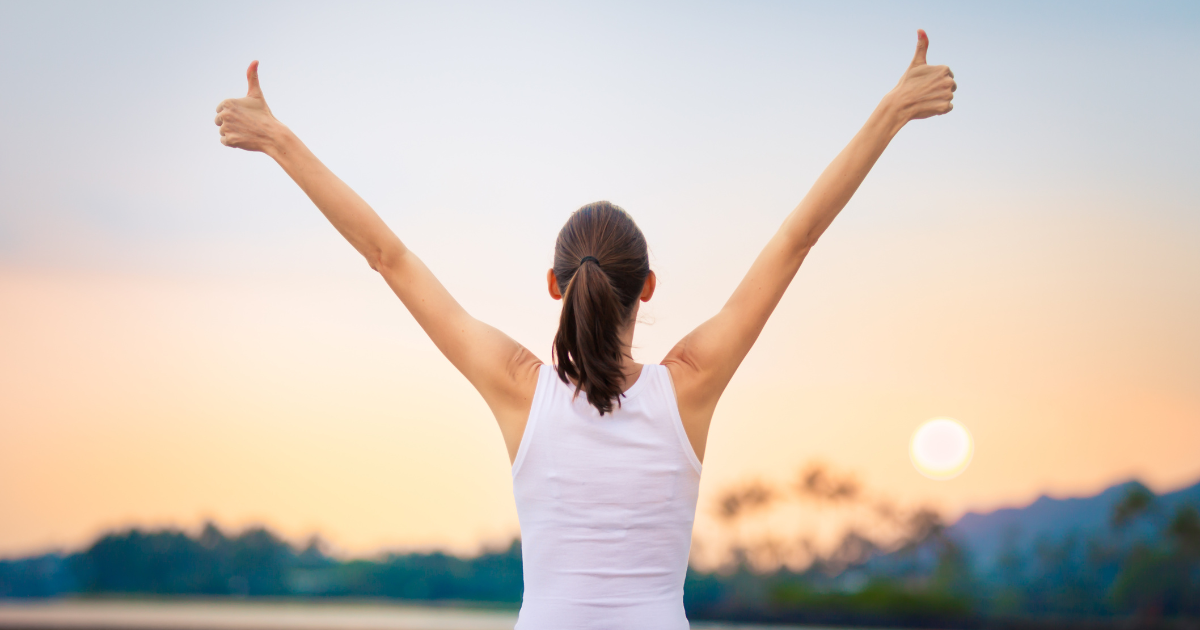 A woman stands outdoors at sunrise or sunset with her back to the camera, arms raised in a V shape, giving two thumbs up. She wears a white tank top, and the sky is soft with pastel colors.