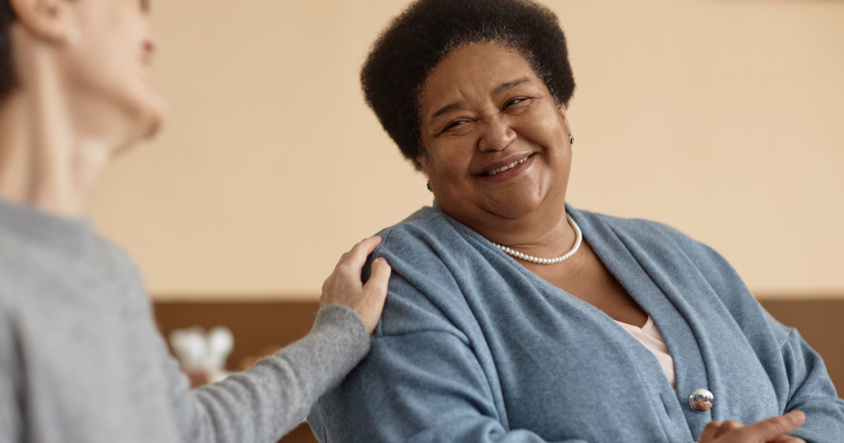 A smiling woman in a blue cardigan sits while another person gently touches her shoulder, both appearing engaged in a warm and supportive conversation about how to support someone in psychiatric treatment.