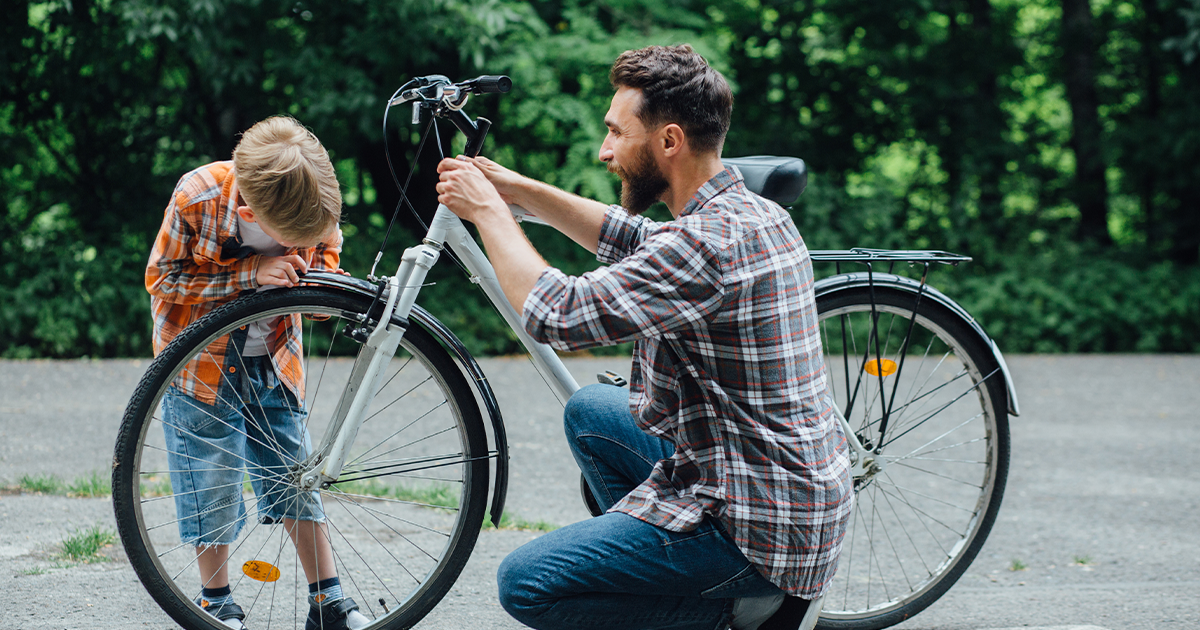 A man kneels, fixing the front wheel of a bicycle as a young boy leans over to watch closely. Both, dressed in plaid shirts and surrounded by green trees, share a moment of connection reminiscent of personalized mental health care outdoors.