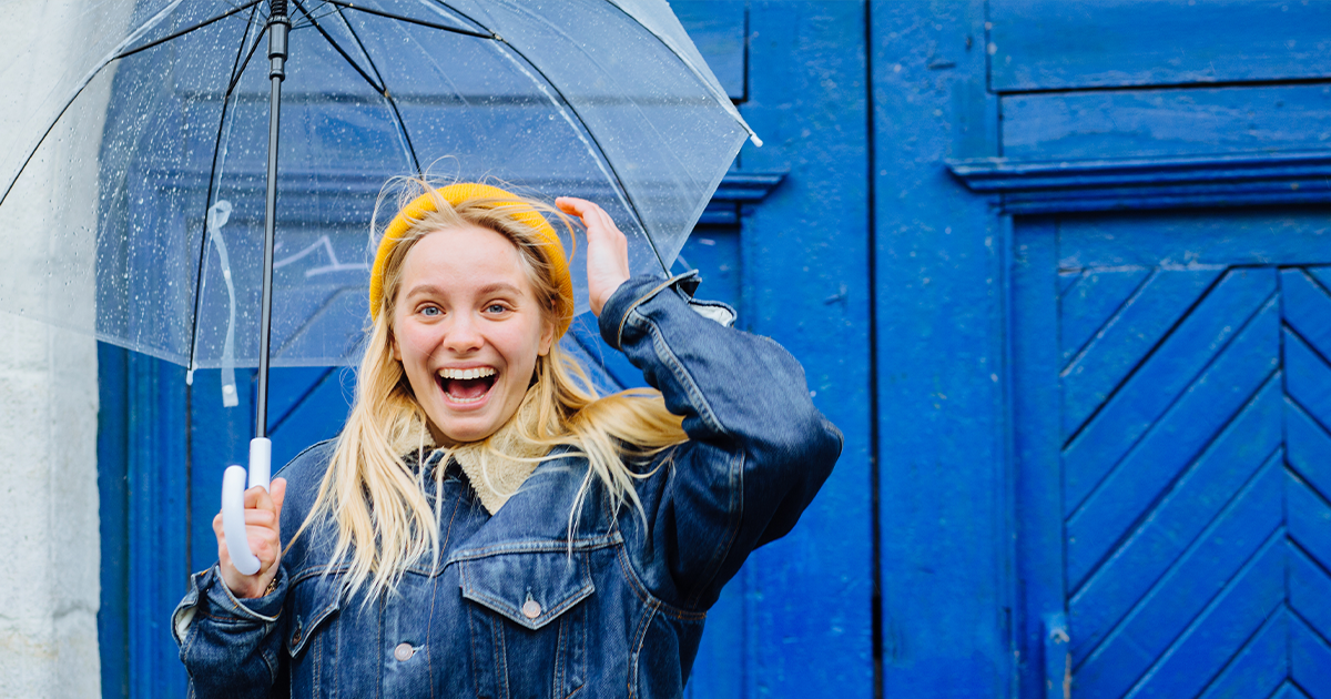 A smiling woman with long blonde hair, wearing a yellow headband and denim jacket, holds a transparent umbrella in front of a bright blue wooden door, challenging psychiatric care myths with her confident presence.