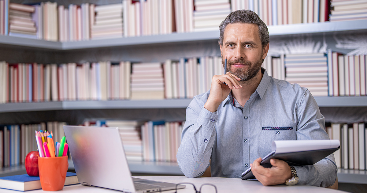 A man with a beard sits at a desk in front of a laptop, holding a notebook and pen. Shelves filled with books are in the background, and psychiatric medication is visible beside a cup of colored pencils and an apple on the desk. He is looking at the camera.