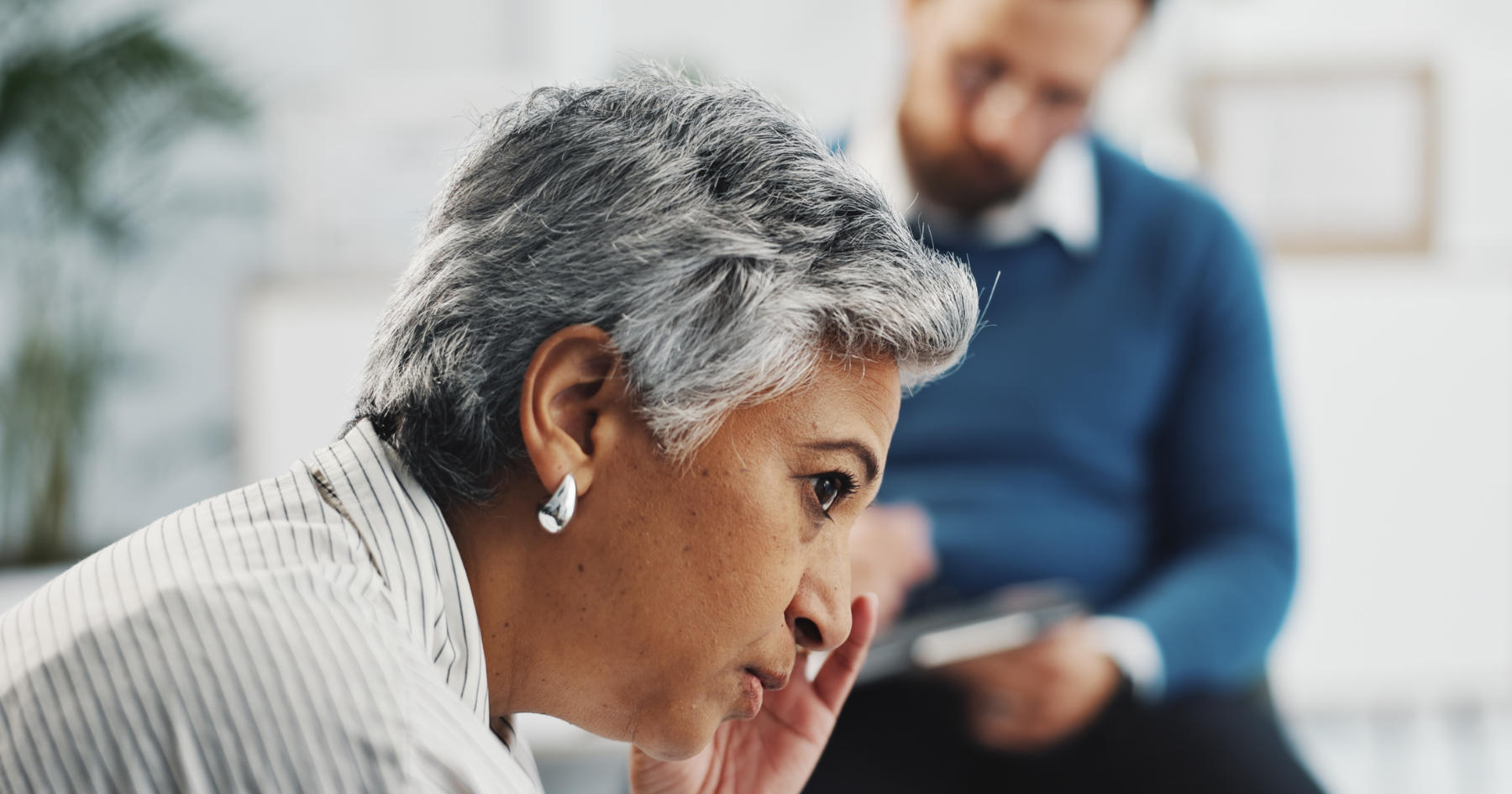 A woman with short gray hair looks thoughtful while sitting indoors; in the blurred background, a person in a blue sweater appears to be writing on a clipboard.