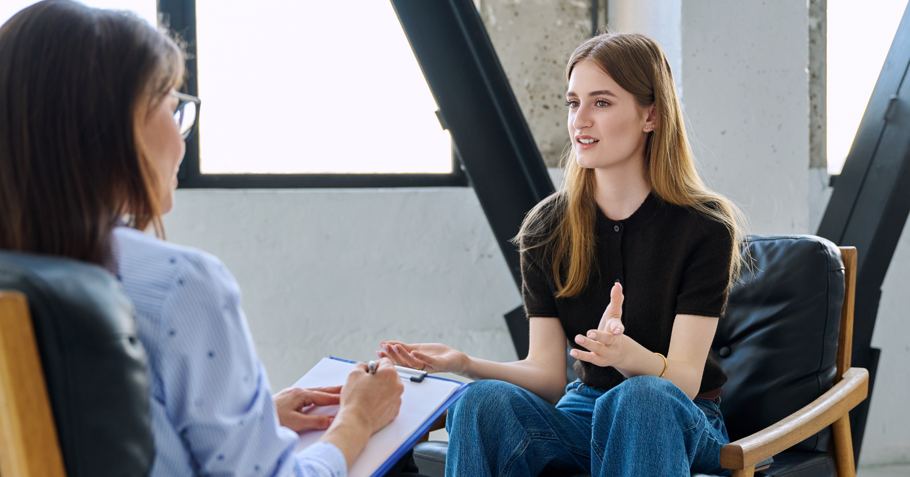 A young woman sits on a chair, talking and gesturing with her hands, while another person holding a clipboard listens and takes notes in a bright, modern office setting.