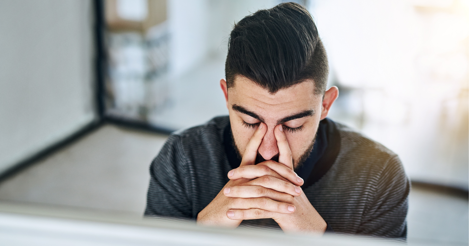 A man with dark hair sits at a desk, eyes closed and pinching the bridge of his nose with both hands, appearing stressed or fatigued in an office setting.