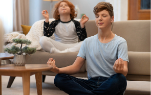 Two children sit cross-legged indoors, meditating with eyes closed and hands in a mudra gesture—a peaceful scene reflecting mindfulness as part of mental health treatment. One sits on the floor, the other relaxed on a couch behind them.