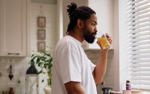 A man with a beard and dreadlocks, wearing a white t-shirt, stands in a kitchen by a window with blinds, sipping orange juice as he takes a moment to manage his anxiety.