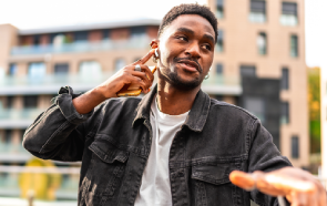 A man wearing a black denim jacket and white shirt smiles while touching his wireless earbud, holding a phone, and gesturing with his other hand outdoors, demonstrating positive coping skills in front of a modern building.