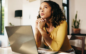 A woman with curly hair, wearing a yellow top, sits at a table with a laptop, resting her chin on her hands and looking thoughtfully upward. Sunlight streams in as she contemplates burnout and depression.