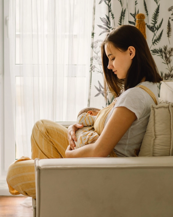 A woman sits on a couch in a softly lit room, breastfeeding her baby. She looks down at the infant, cradling them gently, with leafy-patterned curtains in the background.