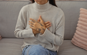 A person wearing a light gray turtleneck sweater and blue jeans sits on a couch with hands crossed over their chest, displaying tattoos on their fingers—a calm moment that reflects the serenity holistic psychiatric care can bring. Pink pillows are visible in the background.