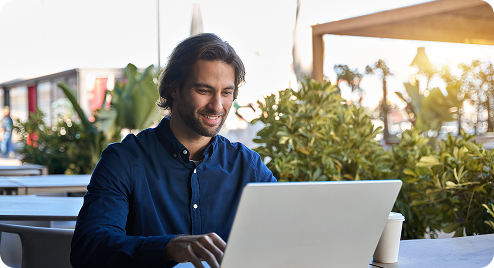 Smiling man working on a laptop at an outdoor café table with plants in the background