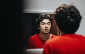 A young person with curly dark hair and a red shirt looks at their reflection in a bathroom mirror, appearing thoughtful or serious, perhaps contemplating how stress affects mental health.