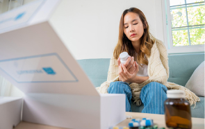 A woman sits on a couch, wrapped in a blanket, looking intently at a medication bottle. An open box and various medicine bottles and blister packs are spread across the table in front of her.