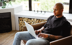 A person sits in a modern chair by a window, using a laptop on their lap to research psychiatric medication help. They wear a black shirt and jeans, with stacked firewood visible in the background.