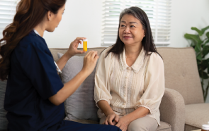 A nurse in navy scrubs explains a bottle of starting psychiatric medication to an older woman sitting on a beige sofa, who is listening and smiling. They are in a bright, home-like setting with natural light.