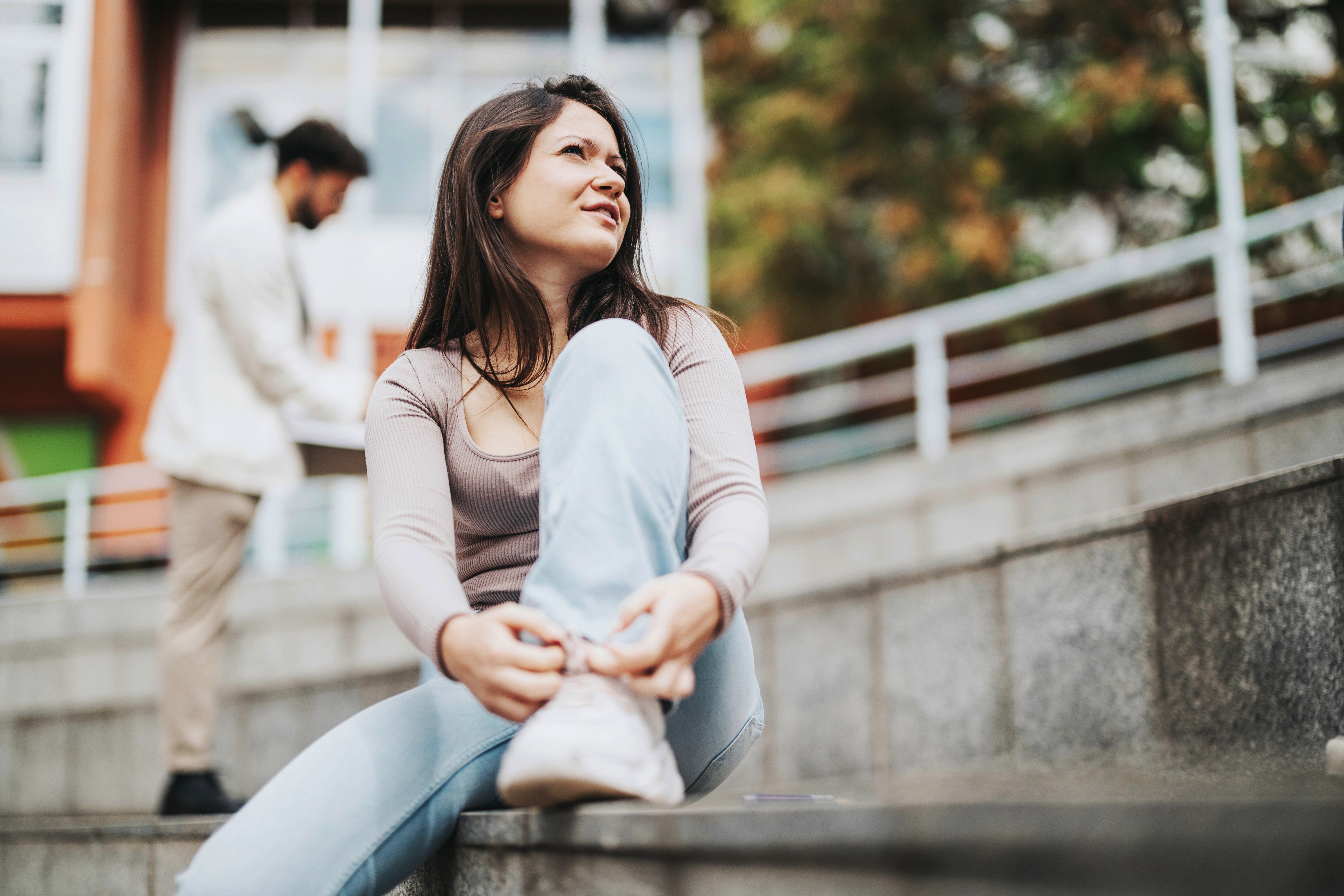 Young woman sitting on outdoor steps holding her ankle with a pained expression