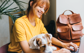 A woman in a yellow shirt smiles while working on her laptop at a table, perhaps researching therapy and medication. She has a small dog on her lap, and a brown leather backpack sits on the table beside her.