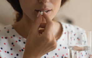 A woman holding a glass of water in one hand prepares to take a white pill with the other, practicing careful medication management. Only the lower half of her face is visible. She wears a white shirt with red and blue polka dots.